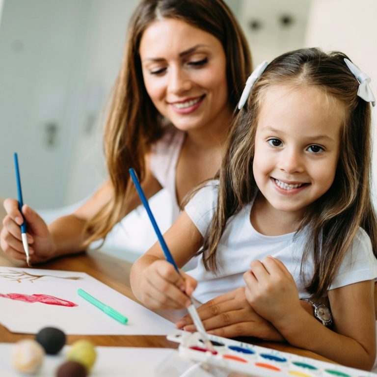 Woman and girl painting together, smiling, with art supplies on a table. Educated Governesses.