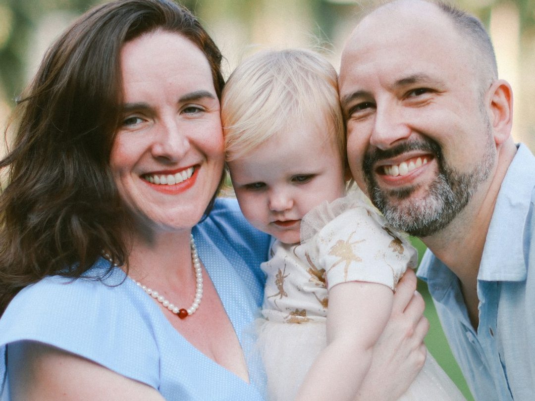 A family portrait featuring a woman, a man, and a toddler outdoors, smiling together.
