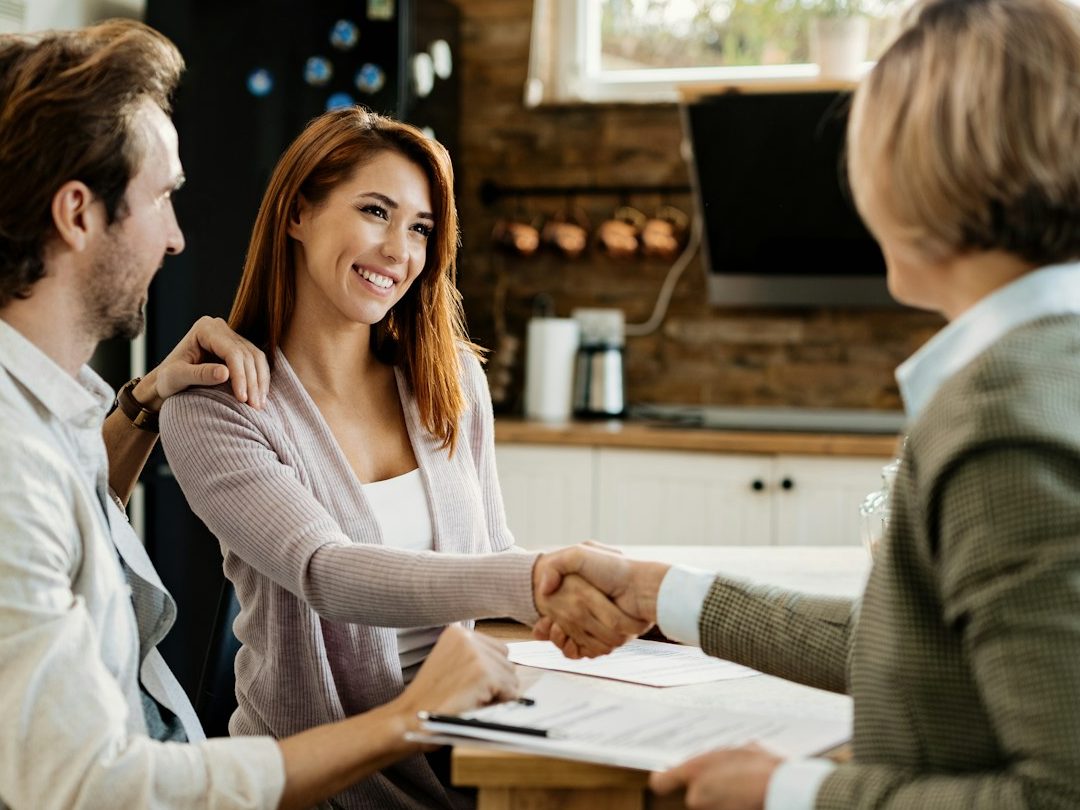 A woman shakes hands with a couple in a modern kitchen setting. Nanny Consulting Services for Families in London, UK and Overseas.